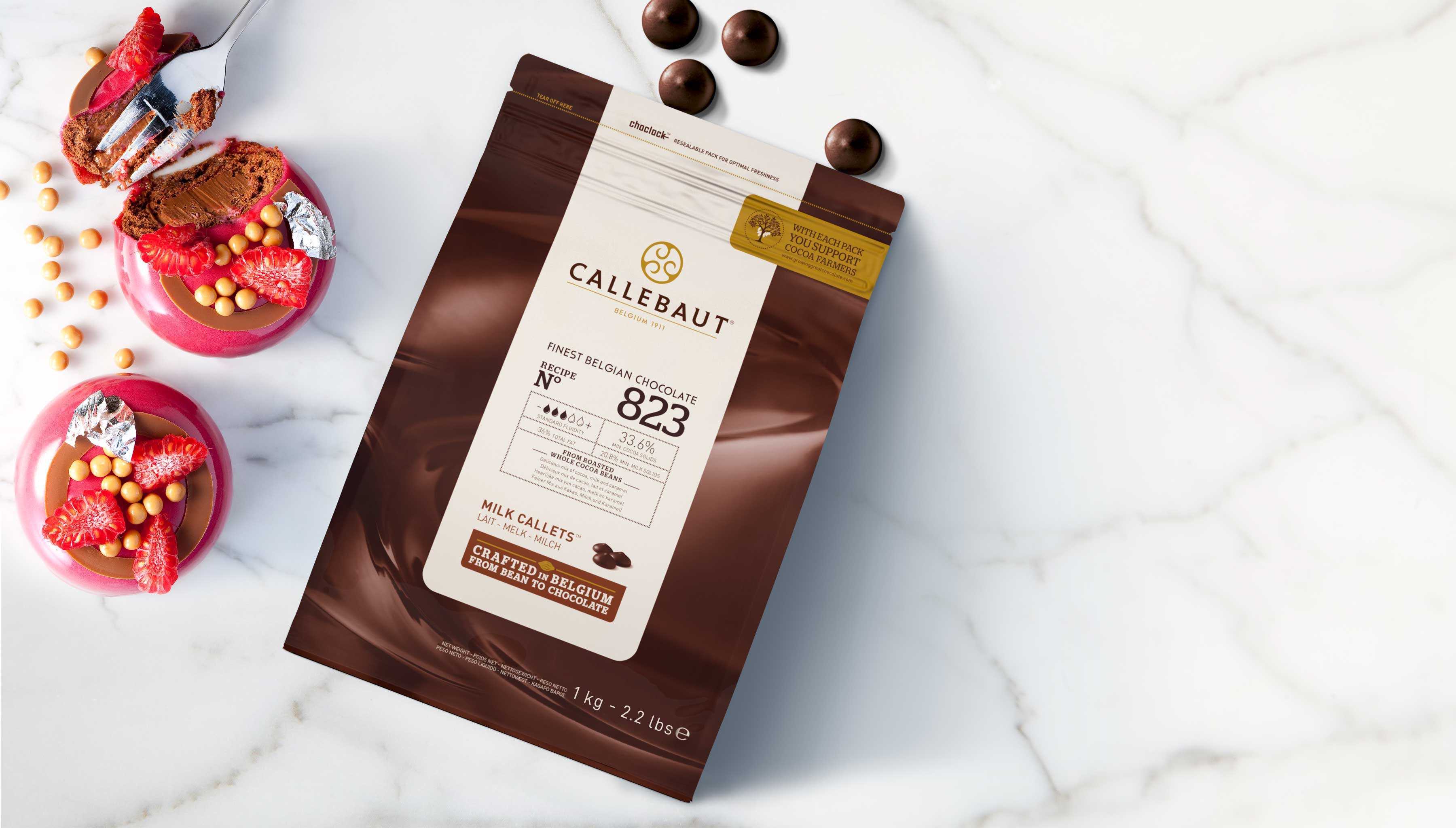 Callebaut Online-Shop – Belgische Kuvertüre und Schokoladen-Callets für Profis. Verpackung mit Callebaut Milchschokolade-Callets auf Marmor, umgeben von Schokostücken und Erdbeeren.